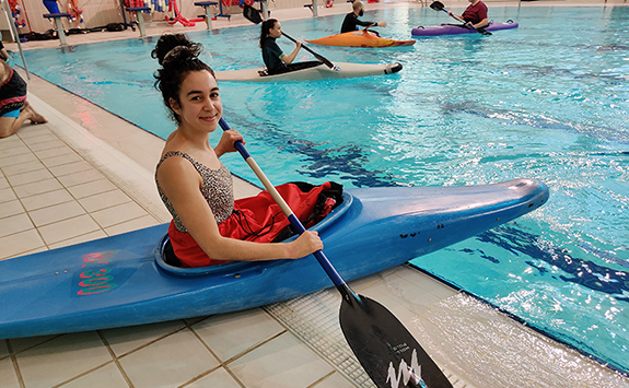 Woman sitting in a blue kayak at the edge of an indoor swimming pool, holding an oar